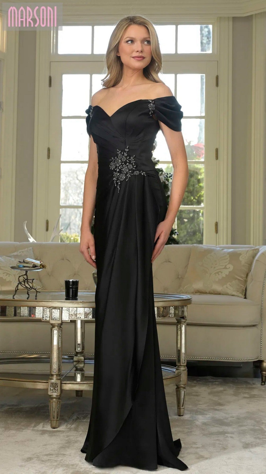 A woman stands indoors in a black Marsoni MV1351 long formal dress with floral embellishments, posed in front of a sofa and large window.